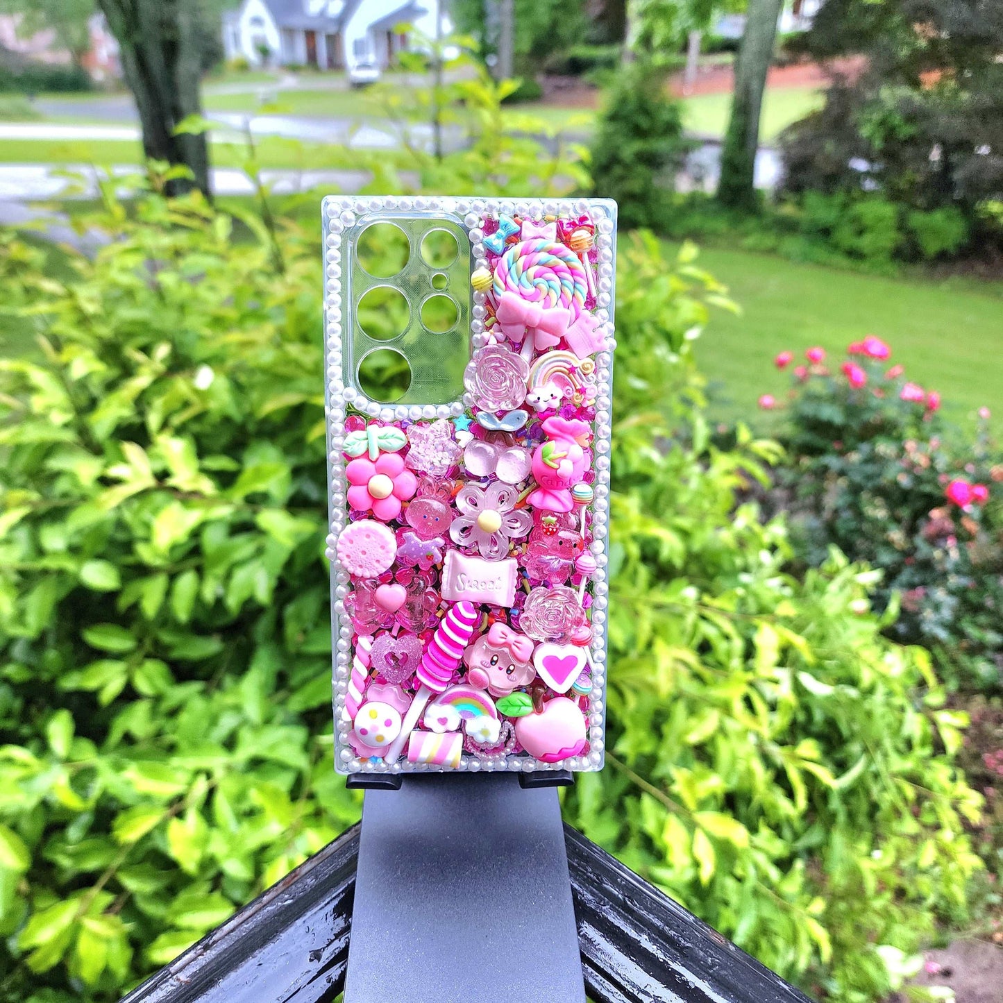 Handmade Kawaii Pink Candy Decoden Phone Case with colorful charms and sparkling designs, compatible with iPhone and Samsung, displayed outdoors.